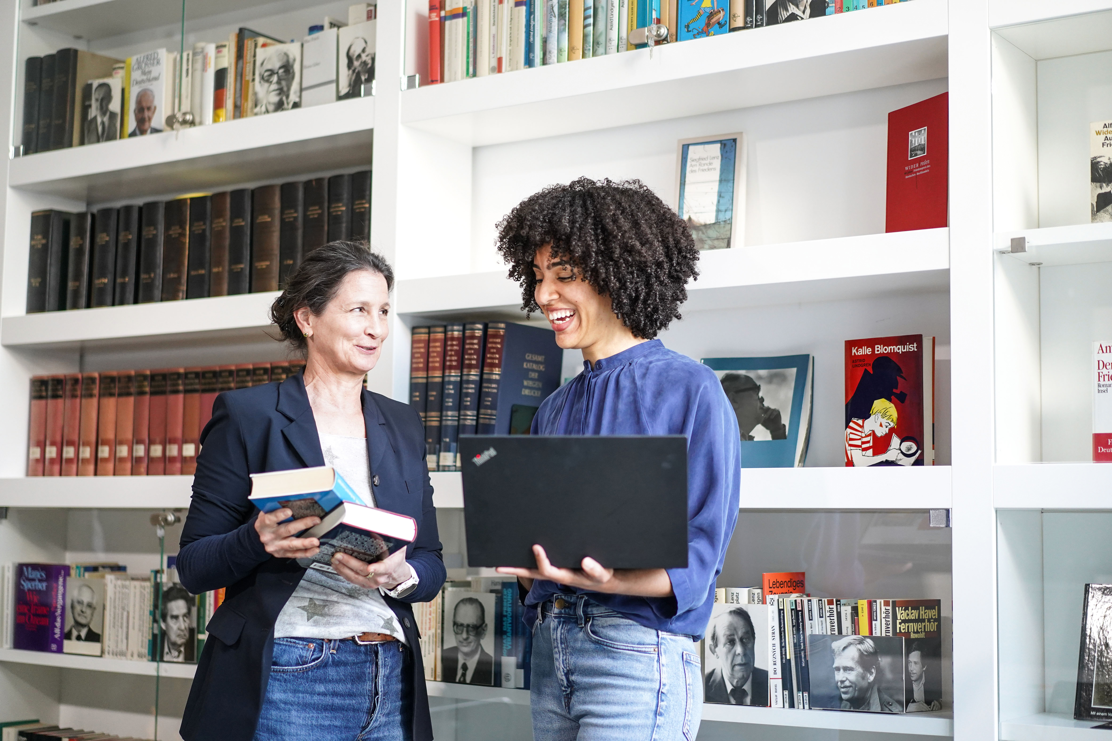 Eine Frau hält Bücher in der Hand, eine weitere einen Laptop. Beide unterhalten sich.