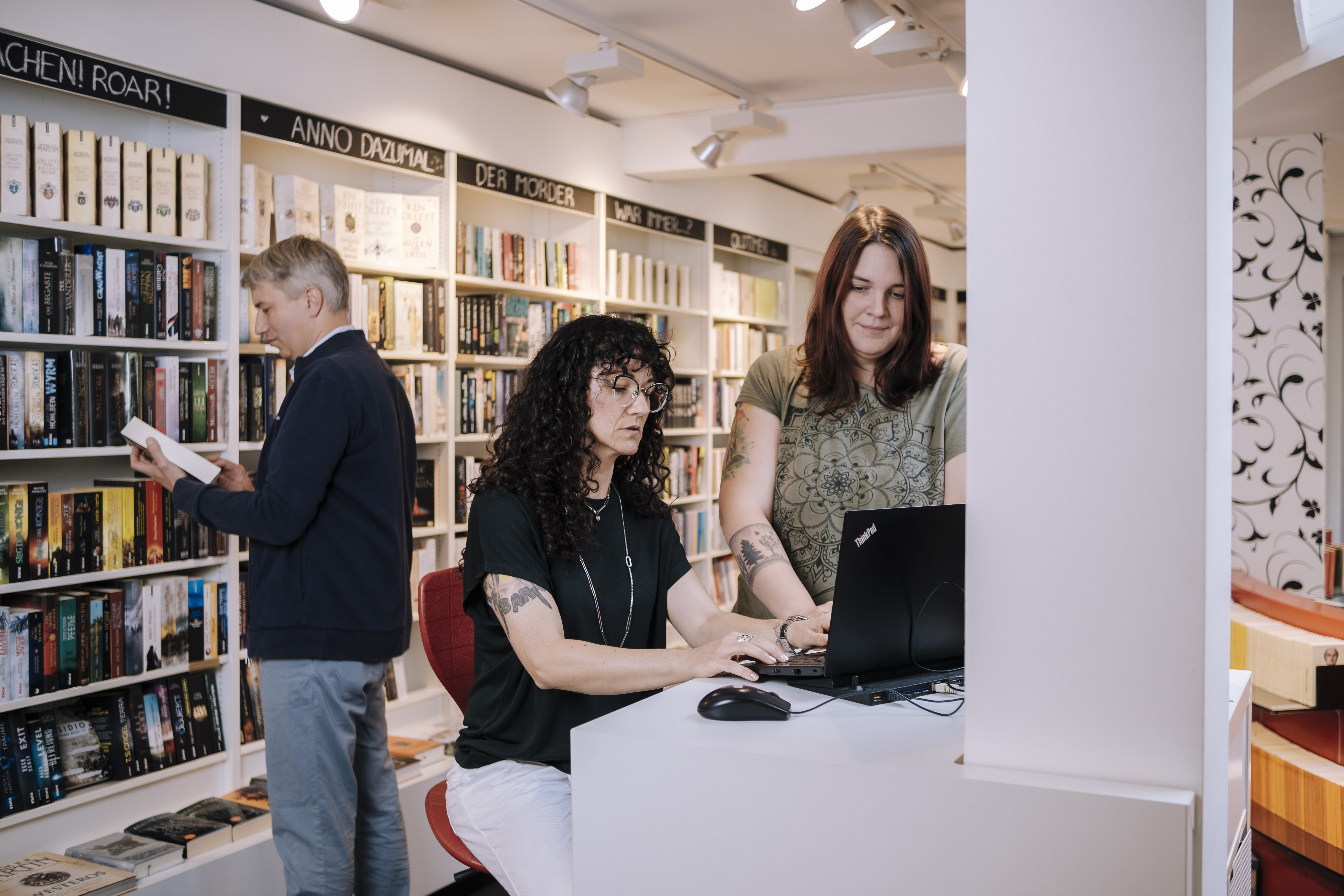 Ein Mann steht an einem Bücherregal in einer Buchhandlung. Im Vordergrund sind zwei Frauen, die sich unterhalten und auf einen Laptop schauen.