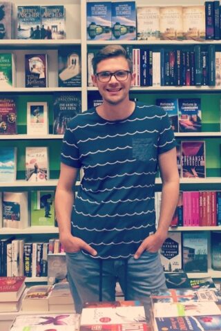 Florian Valerius steht vor einem Bücherregal und lächelt.