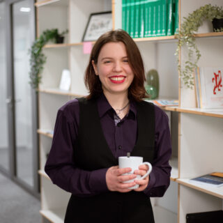 Portrait von Anna Göckel, die vor einem Bücherregal steht, eine Tasse in der Hand hält und lächelt.