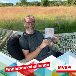 Markus Köcher sitzt in einer Hängematte und hält im Rahmen der Indiebookchallenge im August das Buch "Mucho Mojo" in den Händen und lächelt.