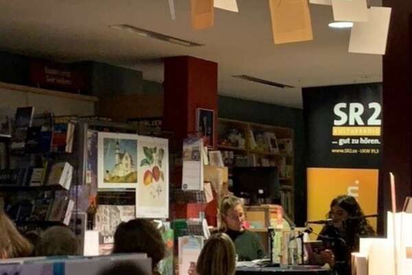 Blick über Bücher hinweg auf eine Autorin, die in einer Buchhandlung ihr Buch vorstellt.