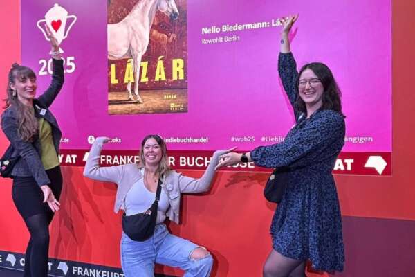 Rahel, Alicia und Franzi zeigen auf einen großen Bildschirm, auf dem das Buch Lazar von Nelio Biedermann abgebildet ist