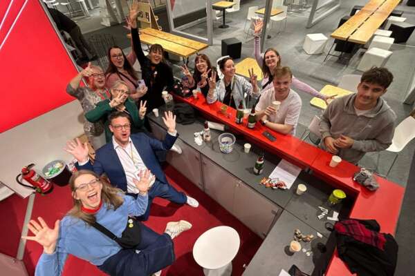 Mehrere Personen stehen und sitzen an der Infotheke, heben die Hände und freuen sich. Das Foto ist von oben aufgenommen