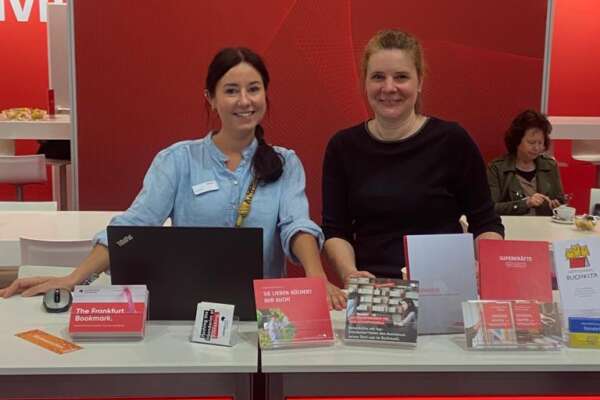 Zwei MVB Mitarbeiterinnen stehen hinter der Theke am MVB Stand und lächeln.