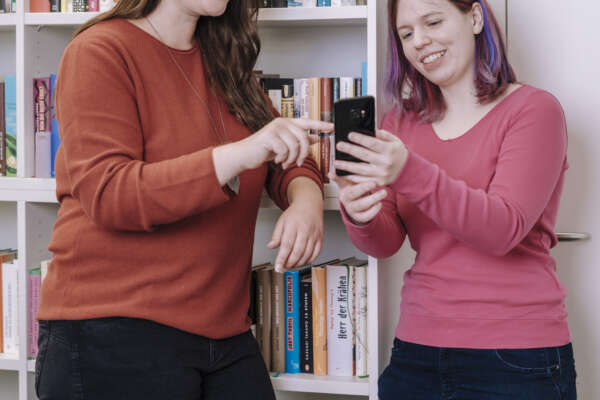 Zwei Frauen stehen vor einem Bücherregal und schauen gemeinsam in ein Handy.