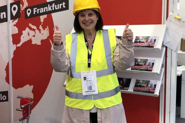 Eine Mitarbeiterin von MVB UK steht in Baustellen Schutzkleidung vor dem MVB Stand auf der London Book Fair, lächelt und streckt zwei Daumen nach oben.