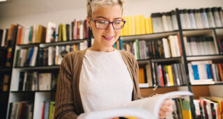 Eine Frau steht vor einem Bücherregal und sieht sich lächelnd ein Buch an.