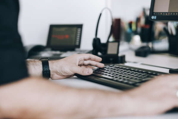 Hände tippen auf eine Tastatur. Im Hintergrund ist eine Arbeitsplatzumgebung.