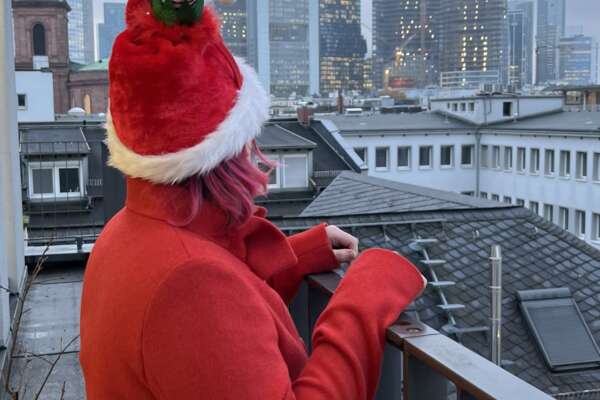 Eine Person steht an einem Geländer und blickt auf die Frankfurter Skyline. Sie trägt einen roten Mantel und eine rote Weihnachtsmütze. 