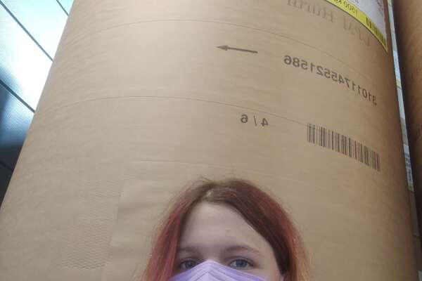 Selfie von Anna, die mit einer Maske im Gesicht vor einer riesigen Papierrolle steht in einer Druckerei. 