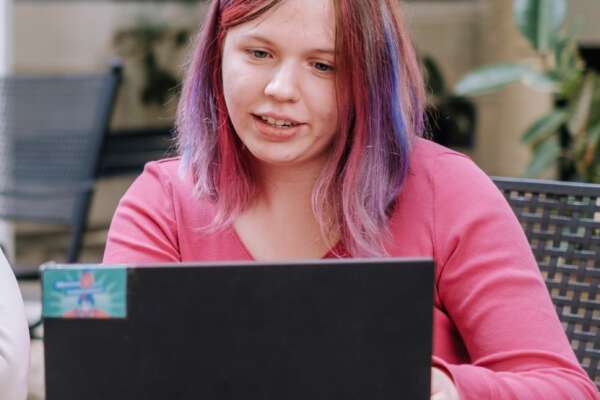 Eine Frau mit rosa Haaren und einem rosafarbenen Oberteil sitzt vor einem Laptop. 