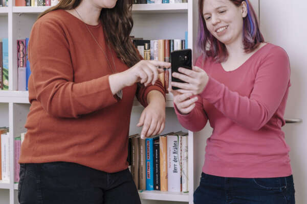 Zwei Frauen stehen vor einem Bücherregal und schauen gemeinsam in ein Handy. 