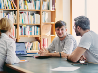 Drei Menschen sitzen an einem Tisch vor einem Bücherregal und schauen auf einen Laptop
